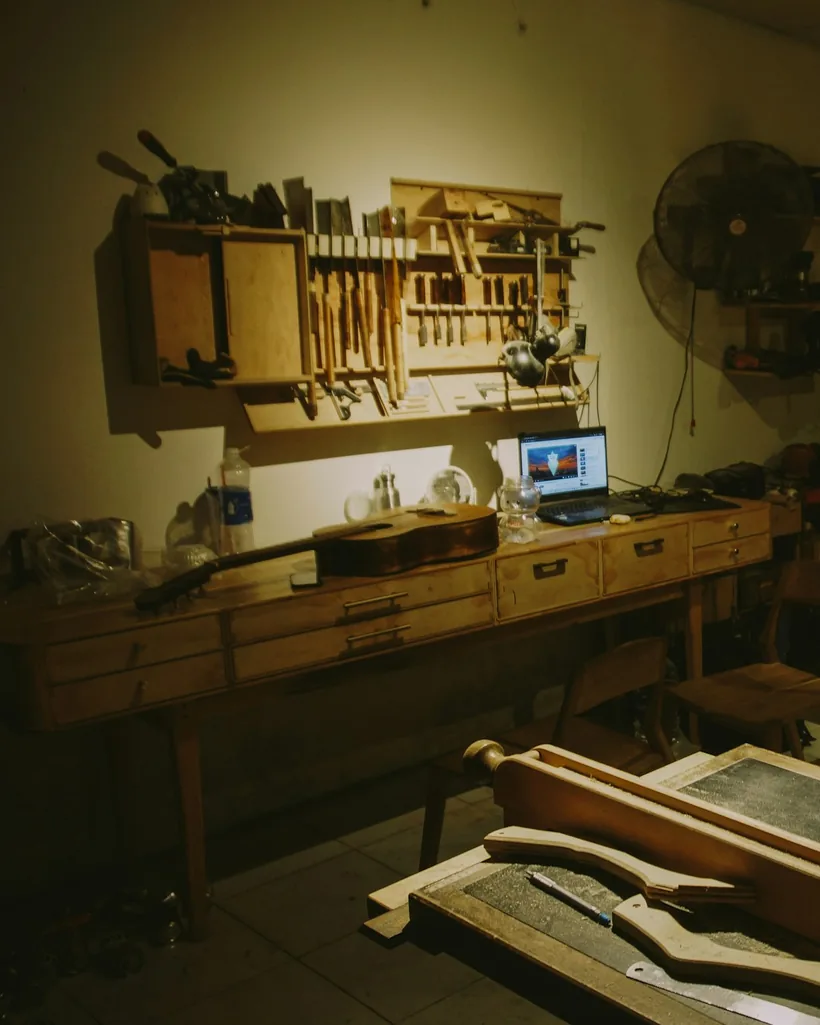 Banco de trabajo de lutería con panel de gubias, formones y cepillos colgados en la pared, y piezas de madera sobre la mesa.
